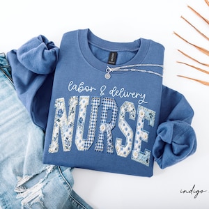 May include: Indigo blue sweatshirt with the words "labor & delivery NURSE" in white script and patchwork letters. The letters are decorated with floral, polka dot, and gingham patterns. A silver necklace rests on the shirt.