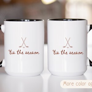 May include: Two white coffee mugs with black handles and rims. Each mug has a brown hockey stick and puck graphic, and the phrase "'tis the season" in brown script. The mugs are displayed on a light surface.