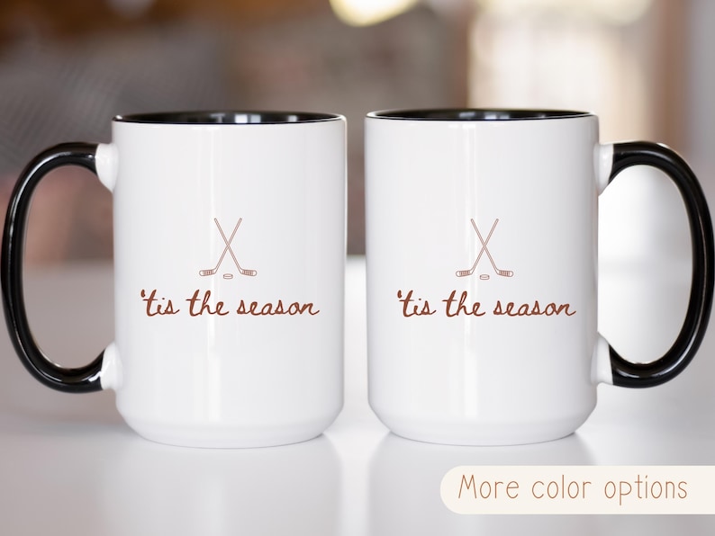 May include: Two white coffee mugs with black handles and rims. Each mug has a brown hockey stick and puck graphic, and the phrase "'tis the season" in brown script. The mugs are displayed on a light surface.