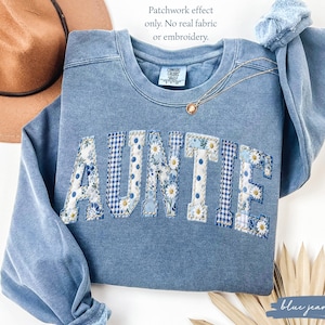 May include: A blue crewneck sweatshirt featuring the word AUNTIE in a printed patchwork effect with floral and gingham patterns. The text Patchwork effect only. No real fabric or embroidery. is visible. The garment has a faded wash finish.