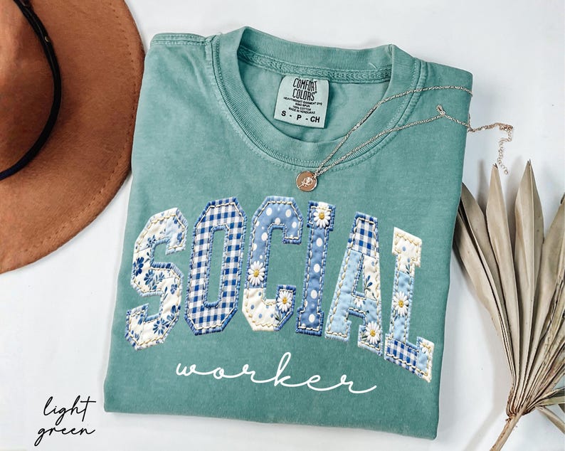 Op de afbeelding: Lichtgroen t-shirt met de woorden "SOCIAL WORKER" in een patchwork-ontwerp. De letters zijn in blauw ruitjes, bloemen- en stippenpatronen. Een gouden ketting met een hanger ligt op het shirt.