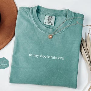 Może przedstawiać: Jasnozielona koszulka z napisem "in my doctorate era" w białym kolorze.