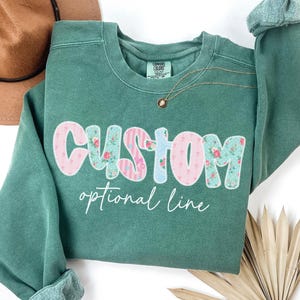 May include: A teal green sweatshirt with the word "CUSTOM" in a floral and striped design, with the words "optional line" below. A gold necklace with a pendant is visible. A brown hat is in the background.