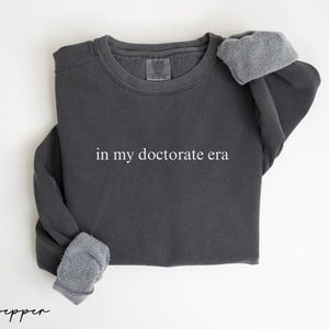 May include: Dark gray sweatshirt with the phrase "in my doctorate era" printed in white. The sweatshirt has a crew neck and ribbed cuffs. The sleeves are folded up, revealing a gray fleece lining. The word "pepper" is written in cursive at the bottom.