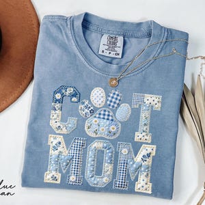 May include: A light blue t-shirt with the words "CAT MOM" appliqued in a patchwork design. The letters and paw print are decorated with floral and gingham patterns in shades of blue and white. A gold necklace with a pendant rests on the shirt.