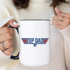 Puede incluir: Taza de cer&aacute;mica blanca con borde y asa negros. La taza presenta el texto "TOP DAD" en azul y rojo, con un gr&aacute;fico de estrella. La taza la sostiene una persona que lleva un su&eacute;ter blanco.