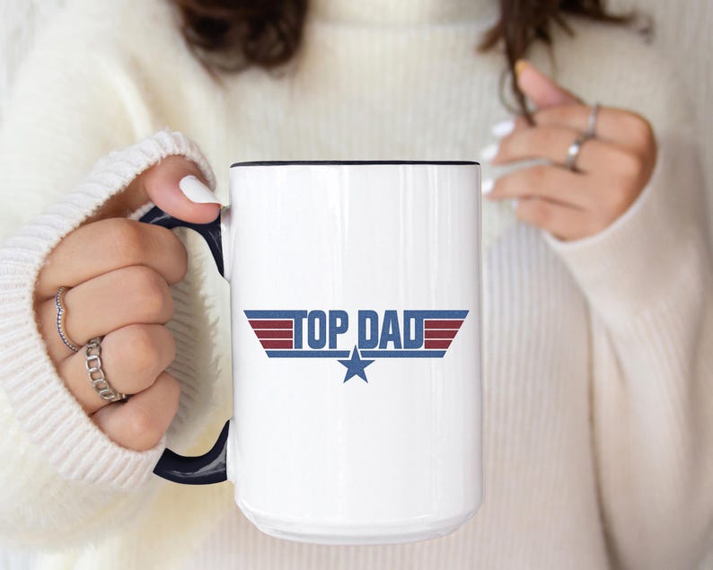 Puede incluir: Taza de cer&aacute;mica blanca con borde y asa negros. La taza presenta el texto "TOP DAD" en azul y rojo, con un gr&aacute;fico de estrella. La taza la sostiene una persona que lleva un su&eacute;ter blanco.