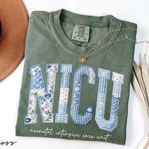 K&ouml;nnte beinhalten: Moosgr&uuml;nes T-Shirt mit den Buchstaben "NICU", die mit blauen Blumen-, Tupfen- und Vichy-Mustern appliziert sind. Darunter steht der Schriftzug "neonatal intensive care unit". Eine goldene Halskette und ein brauner Hut sind ebenfalls zu sehen.