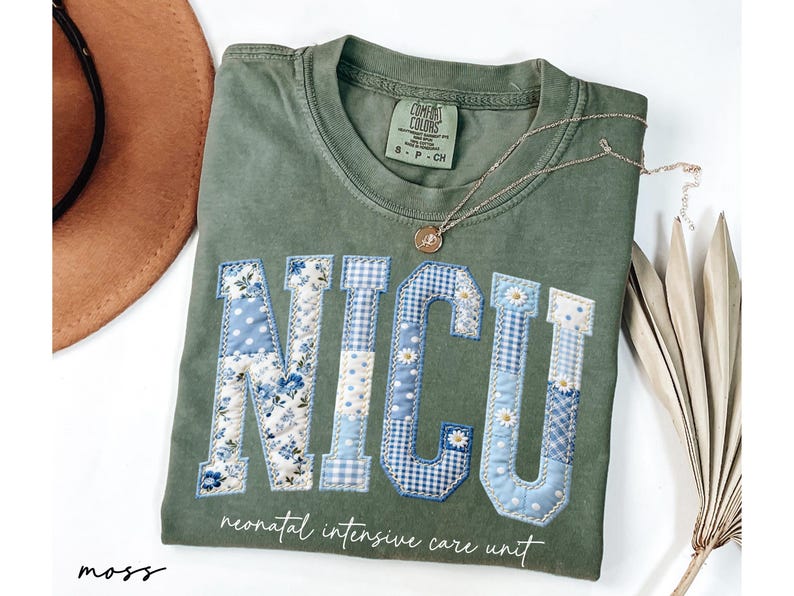 K&ouml;nnte beinhalten: Moosgr&uuml;nes T-Shirt mit den Buchstaben "NICU", die mit blauen Blumen-, Tupfen- und Vichy-Mustern appliziert sind. Darunter steht der Schriftzug "neonatal intensive care unit". Eine goldene Halskette und ein brauner Hut sind ebenfalls zu sehen.