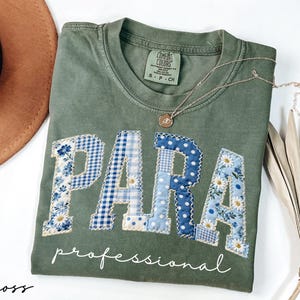 May include: Moss green t-shirt with the word "PARA" in patchwork letters, featuring blue floral and gingham patterns. The word "professional" is written below in white script. A gold necklace with a pendant rests on the shirt.