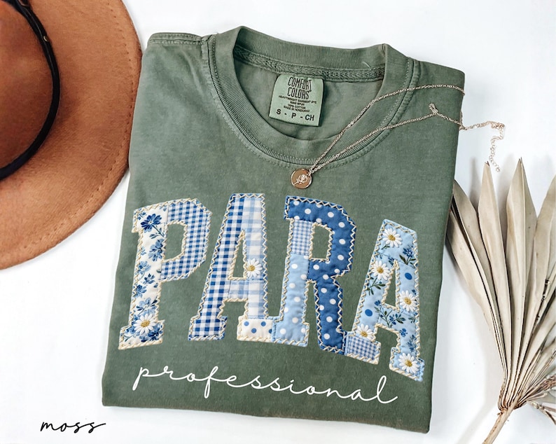 May include: Moss green t-shirt with the word "PARA" in patchwork letters, featuring blue floral and gingham patterns. The word "professional" is written below in white script. A gold necklace with a pendant rests on the shirt.