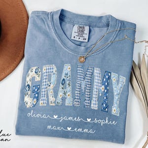 May include: A blue jean Comfort Colors t-shirt with the word "Grammy" in patchwork letters. The letters are decorated with floral and gingham patterns. Below the word are names: Olivia, James, Sophie, Max, and Emma. A gold necklace rests on the shirt.