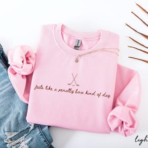 Op de afbeelding: Lichtroze sweatshirt met de tekst "feels like a penalty box kind of day" en gekruiste hockeysticks. Een gouden ketting met een hanger ligt op de trui. Het sweatshirt wordt gecombineerd met een blauwe jeans.