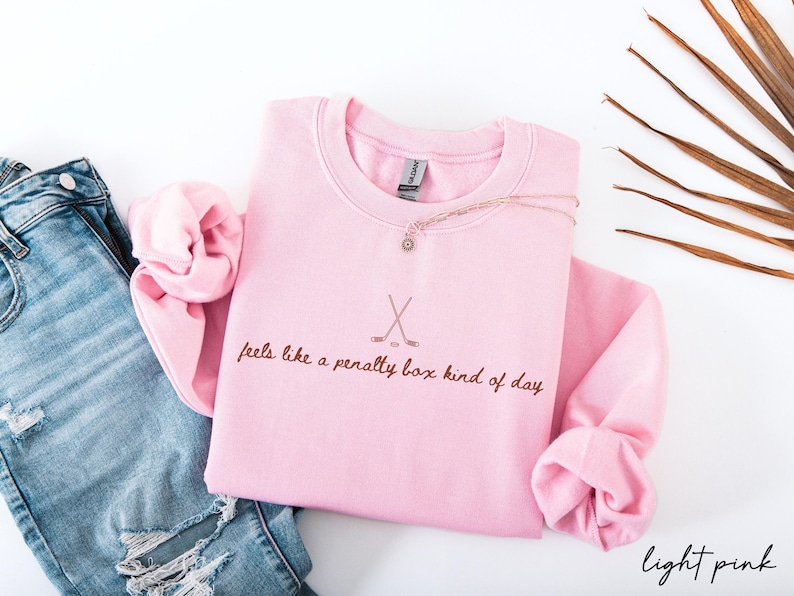 Op de afbeelding: Lichtroze sweatshirt met de tekst "feels like a penalty box kind of day" en gekruiste hockeysticks. Een gouden ketting met een hanger ligt op de trui. Het sweatshirt wordt gecombineerd met een blauwe jeans.
