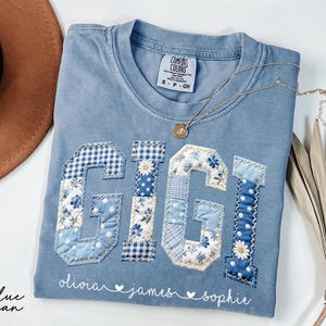 May include: A blue jean Comfort Colors t-shirt with the name "GIGI" appliqued in patchwork fabric. The letters feature floral, gingham, and polka dot patterns. The names "olivia", "james", and "sophie" are embroidered below. A gold necklace rests on the shirt.