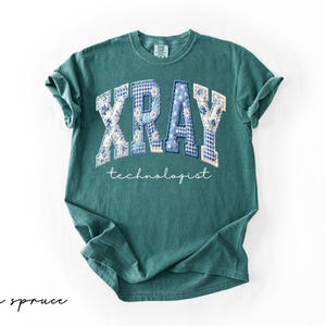May include: A teal-colored t-shirt with the word "XRAY" in a patchwork design of blue floral and gingham patterns. Below, the word "technologist" is written in a delicate script. The shirt is a comfortable, casual style.