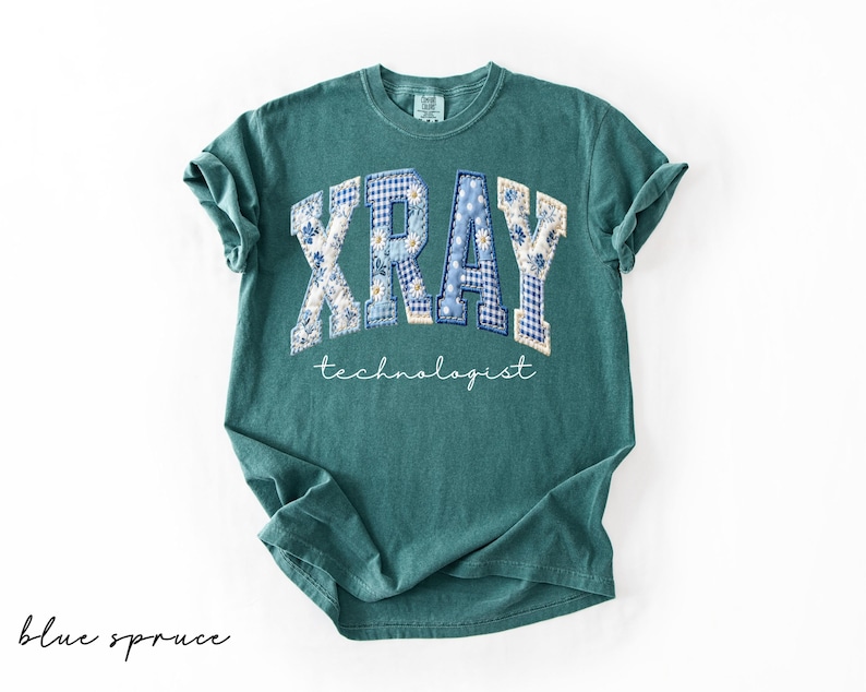 May include: A teal-colored t-shirt with the word "XRAY" in a patchwork design of blue floral and gingham patterns. Below, the word "technologist" is written in a delicate script. The shirt is a comfortable, casual style.