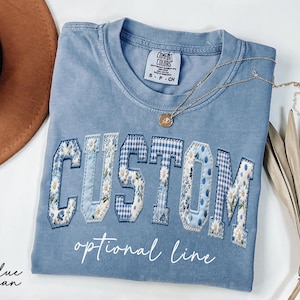 May include: A blue jean Comfort Colors t-shirt with the word "CUSTOM" in a patchwork design of floral and gingham patterns. The shirt also features the text "optional line" in a script font. A gold necklace with a pendant rests on the shirt.