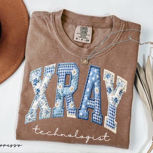 May include: Espresso-colored t-shirt with the word "XRAY" in a patchwork design featuring blue floral and gingham patterns. The word "technologist" is written below. A silver necklace with a small pendant is draped across the shirt.