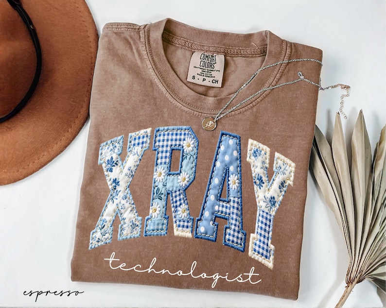 May include: Espresso-colored t-shirt with the word "XRAY" in a patchwork design featuring blue floral and gingham patterns. The word "technologist" is written below. A silver necklace with a small pendant is draped across the shirt.