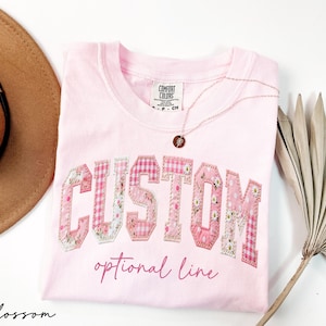 May include: A light pink t-shirt with the word "CUSTOM" in a patchwork design, featuring floral and gingham patterns. The text "optional line" is below the word "CUSTOM". A gold necklace and a brown hat are also in the image.
