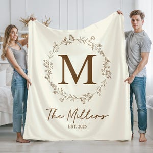 May include: Cream-colored blanket featuring a large brown letter "M" inside a floral wreath. Below the letter is the text "The Millers" and "EST. 2025" in a brown script. The blanket is held up by two people.