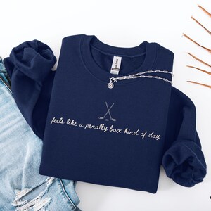 Op de afbeelding: Marineblauwe sweatshirt met witte borduursels met de tekst "feels like a penalty box kind of day". Het ontwerp bevat gekruiste hockeysticks. Een zilveren ketting met een hanger is zichtbaar.