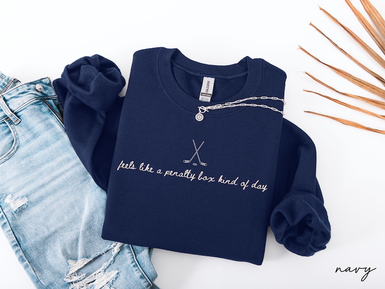 Op de afbeelding: Marineblauwe sweatshirt met witte borduursels met de tekst "feels like a penalty box kind of day". Het ontwerp bevat gekruiste hockeysticks. Een zilveren ketting met een hanger is zichtbaar.