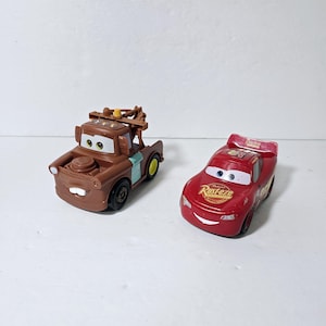 Puede incluir: Dos coches de juguete de la franquicia de películas Cars de Disney Pixar. El primer coche es una grúa marrón con un gancho de remolque amarillo y una franja azul. El segundo coche es un coche de carreras rojo con un número blanco "95" y las palabras "Rust-eze" en el lateral.