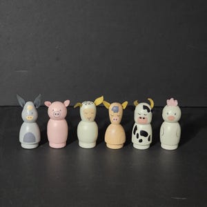 Peut inclure: Ensemble de six figurines d'animaux de ferme en bois. Les figurines sont peintes dans différentes couleurs, notamment le rose, le gris, le blanc, le brun et le noir. Chaque figurine a un visage d'animal différent peint dessus, notamment un cochon, un âne, un mouton, une vache, un poulet et un cheval.