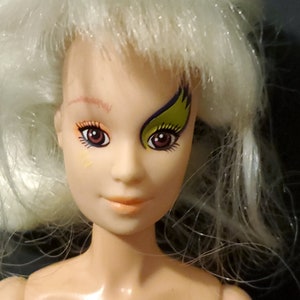 Puede incluir: Una muñeca de pelo blanco con la cara pintada. La muñeca tiene ojos marrones y un diseño amarillo y morado pintado en un ojo.