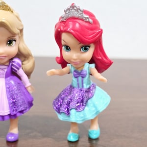 May include: Two small plastic Disney princess dolls. The doll on the left is wearing a purple dress with glitter and a blonde braid. The doll on the right is wearing a blue and purple dress with glitter and a red wig with a silver crown.
