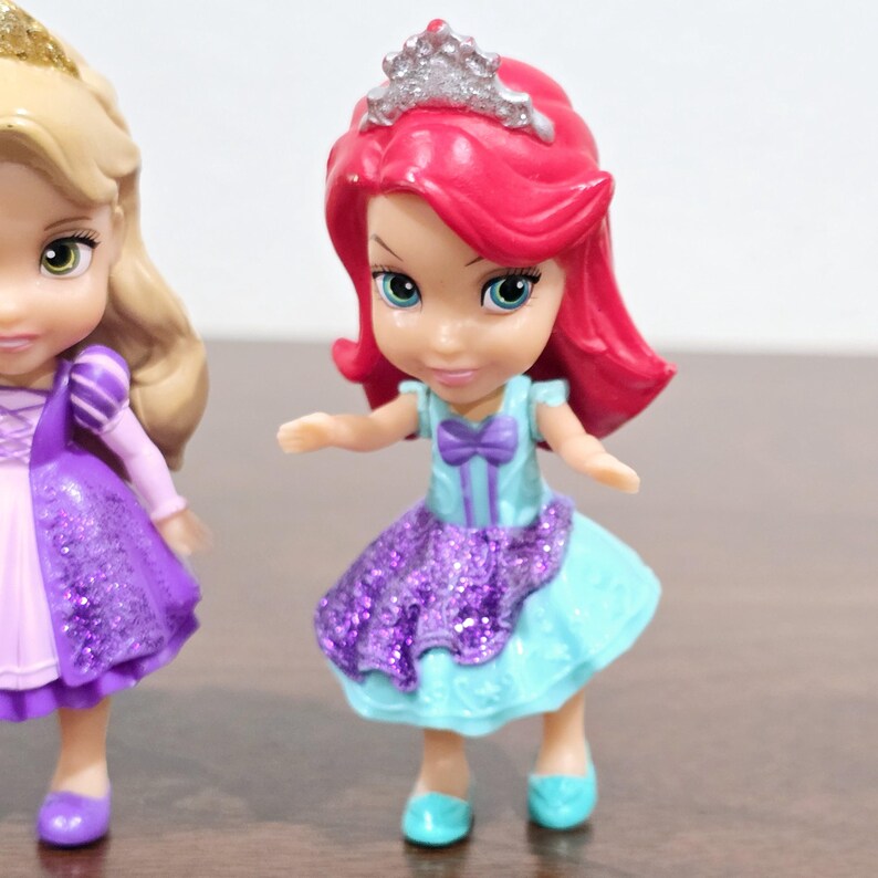 May include: Two small plastic Disney princess dolls. The doll on the left is wearing a purple dress with glitter and a blonde braid. The doll on the right is wearing a blue and purple dress with glitter and a red wig with a silver crown.