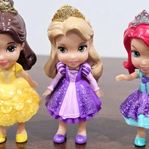 May include: Three small plastic Disney princess dolls. The dolls are wearing sparkly dresses. The princess on the left is wearing a yellow dress, the princess in the middle is wearing a purple dress, and the princess on the right is wearing a blue dress.