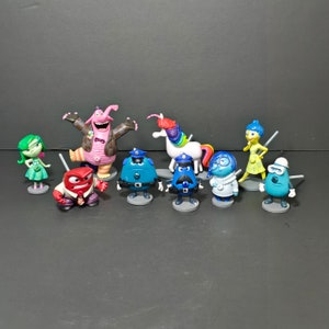 May include: A collection of Inside Out character figurines, including Joy, Sadness, Anger, and Bing Bong. The figures are brightly colored and stand on small gray bases. A white unicorn with a rainbow mane is also present.
