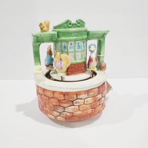 Puede incluir: Un carrusel musical de cerámica con una tienda verde con el texto "Ginger & Pickle" y figuras giratorias de una ardilla, un conejo, una gallina y un pato. El carrusel está decorado con ladrillos y tiene una base blanca.