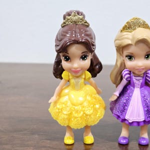 May include: Two small plastic Disney princess dolls. The doll on the left is Belle from Beauty and the Beast, wearing a yellow dress with gold glitter. The doll on the right is Rapunzel from Tangled, wearing a purple dress with gold glitter.
