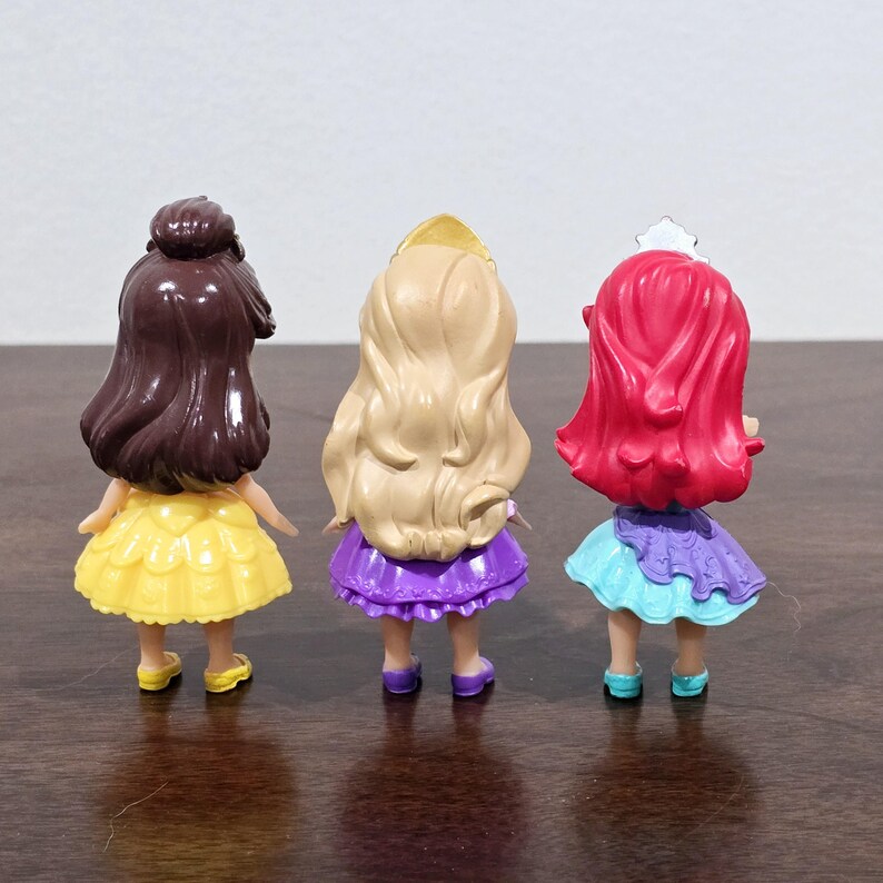 May include: Three small plastic Disney princess dolls. The dolls are wearing dresses in yellow, purple, and blue. The dolls are facing away from the camera.