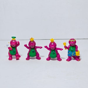 May include: Four purple dinosaur figurines with different accessories. One is wearing a crown and holding a flag that says "GO", another is wearing a crown, another is wearing sunglasses and holding a spade and bucket, and the last one is wearing a crown.