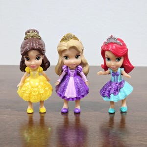 May include: Three small plastic Disney princess dolls. The dolls are Belle, Rapunzel, and Ariel. Belle is wearing a yellow dress with a gold crown. Rapunzel is wearing a purple dress with a gold crown. Ariel is wearing a blue dress with a red crown.