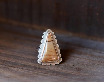 Op de afbeelding: Een zilveren ring met een driehoekige steen met horizontale banden in beige en bruin. De steen is gezet in een zilveren zetting omringd door een decoratieve geschulpte rand met een getextureerde, gestippelde afwerking.