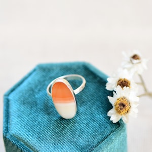 May include: A silver ring featuring an oval-shaped orange and white agate stone set in a silver band. The ring is presented on a teal velvet ring box.