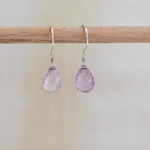 May include: A pair of drop earrings featuring light purple, teardrop-shaped amethyst gemstones carved with a leaf pattern. The stones are suspended from silver-toned metal ear wires, each secured with a small wire-wrapped detail at the top.