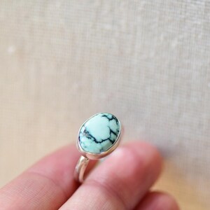 May include: A silver ring with a light blue and black oval gemstone. The ring is on a finger.