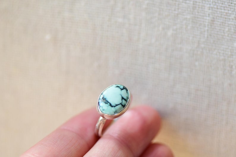May include: A silver ring with a light blue and black oval gemstone. The ring is on a finger.