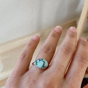 May include: A silver ring with a light blue and black oval gemstone. The ring is on a person's finger.