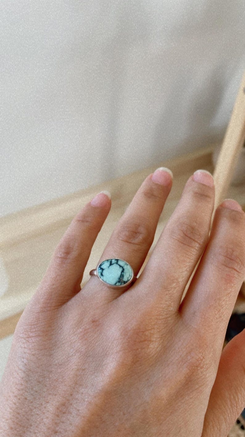 May include: A silver ring with a light blue and black oval gemstone. The ring is on a person's finger.