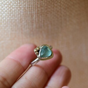 May include: A ring featuring a faceted, triangular light blue gemstone set in a silver bezel with a gold-toned rim. The setting includes a cluster of small gold-toned spheres on one side, attached to a smooth, polished silver band.