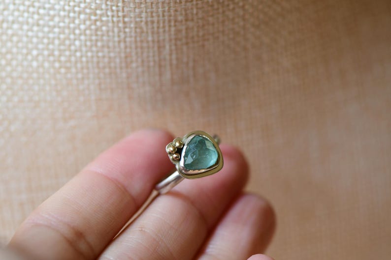 May include: A ring featuring a faceted, triangular light blue gemstone set in a silver bezel with a gold-toned rim. The setting includes a cluster of small gold-toned spheres on one side, attached to a smooth, polished silver band.
