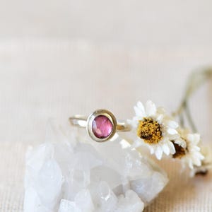 May include: A silver ring with a pink gemstone set in a bezel setting. The ring is resting on a white crystal.